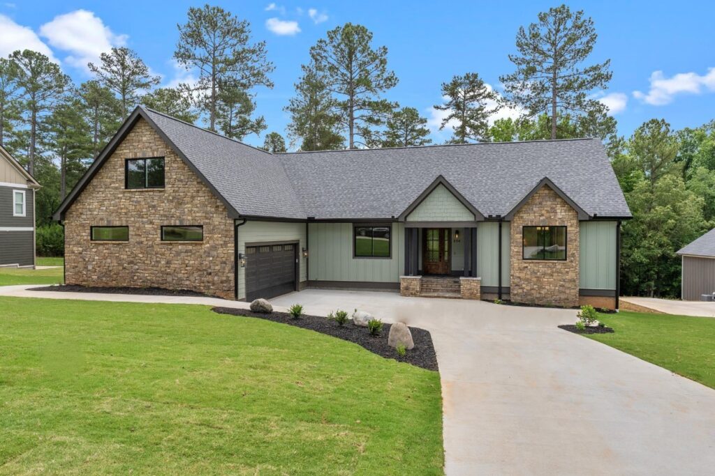 A modern single-story house with stone and green siding, a dark gray roof, a covered front porch, and a two-car garage, surrounded by a well-kept lawn and tall trees in the background.
