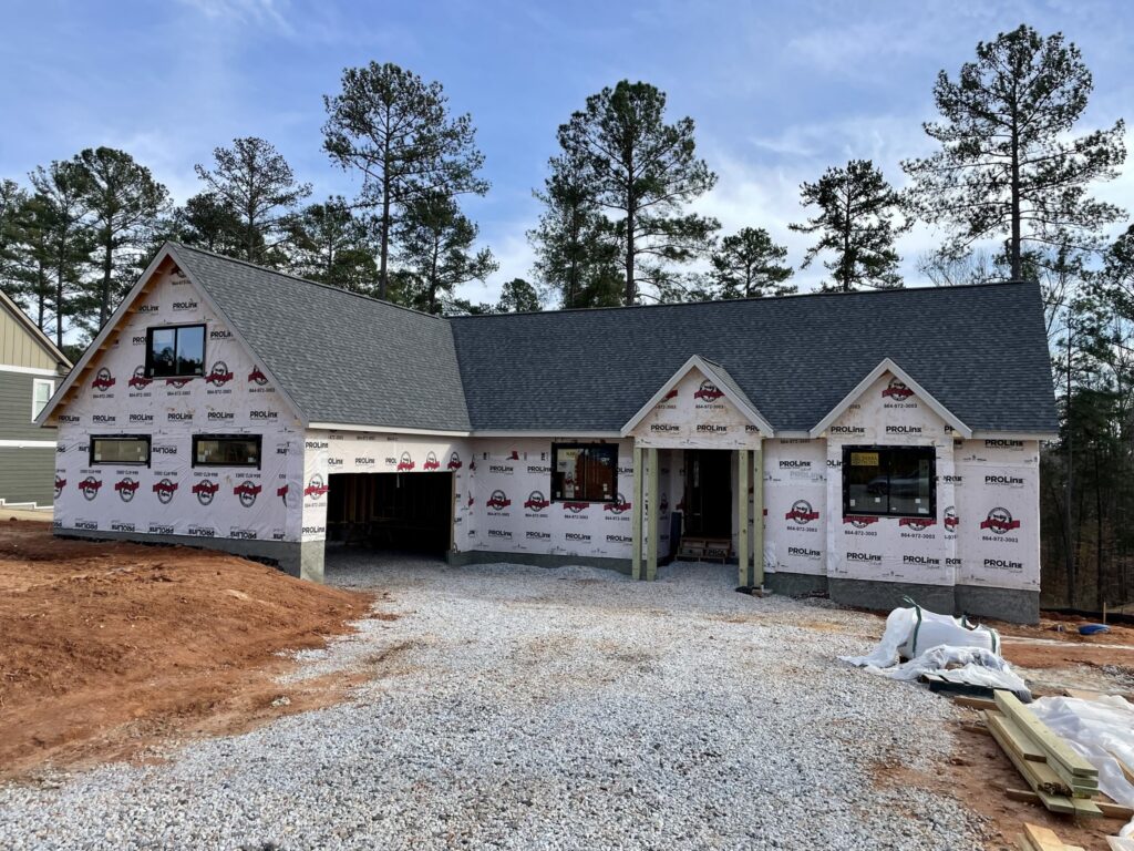 A single-story house under construction, partially covered in white weatherproof sheeting, with a gray shingle roof. The driveway is gravel, and there are trees and building materials in the background.