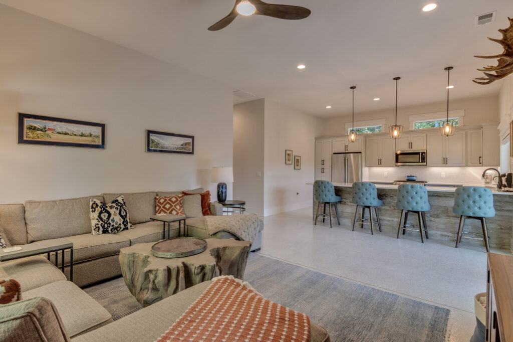 Open-concept living space with a beige sectional sofa, patterned pillows, natural wood coffee tables, and a modern kitchen featuring blue upholstered barstools, pendant lights, and stainless steel appliances.