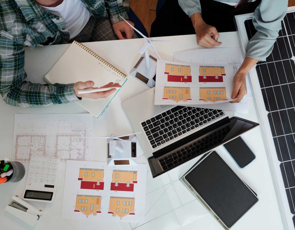 Two people discuss house floor plans at a desk with blueprints, printed layouts, a calculator, a laptop, a tablet, and a solar panel. One person takes notes while the other points to a paper.