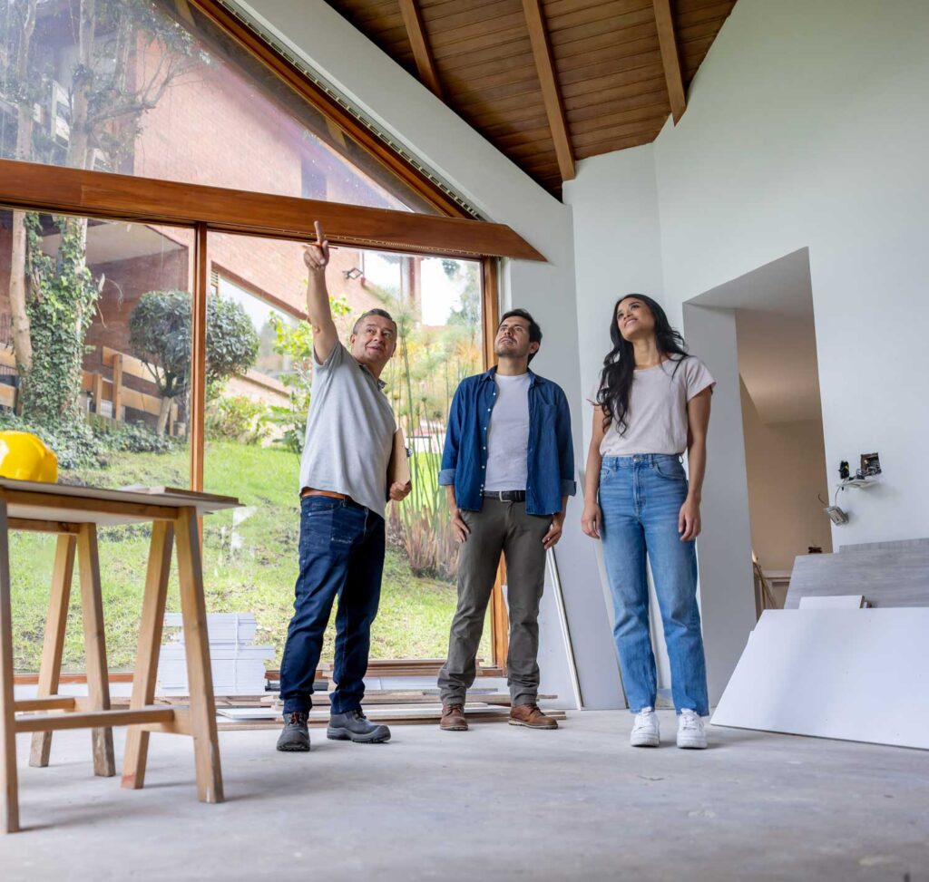 Three people stand in a partially renovated room with large windows. One person is pointing toward the ceiling, while the other two look up attentively. Construction materials and tools are visible on the floor and table.