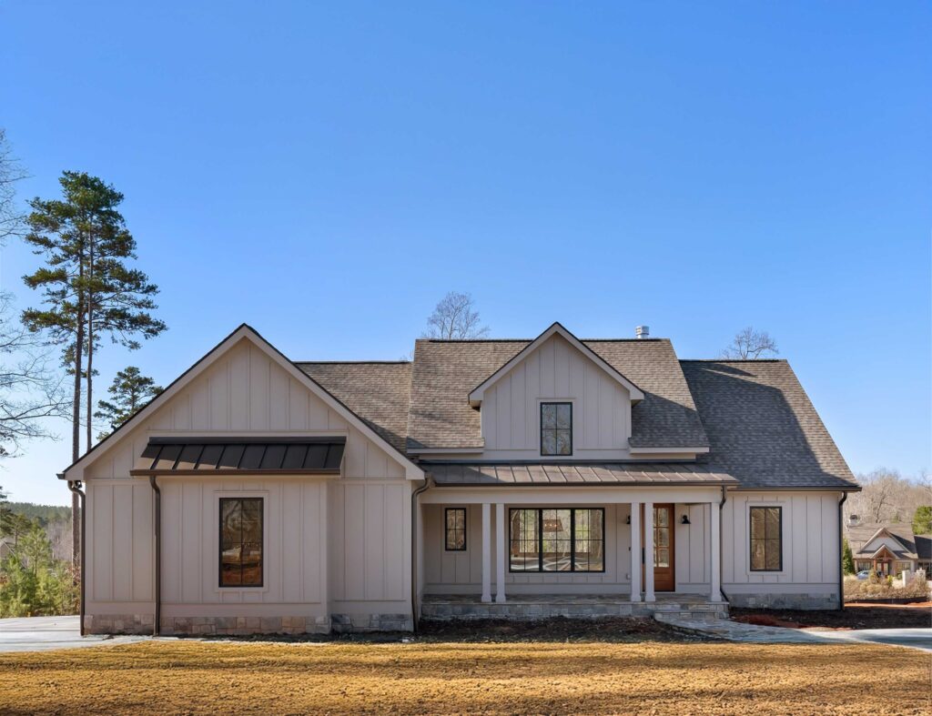 Modern farmhouse-style home with light-colored vertical siding, black-trimmed windows, gabled roof, covered front porch, and a wooden front door, set against a clear blue sky and sparse trees in the background.