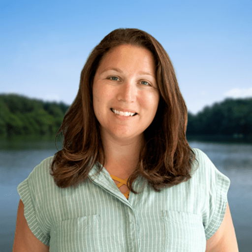 A woman with wavy brown hair and a light green short-sleeve shirt smiles in front of a calm lake with forested hills and a clear blue sky in the background.
