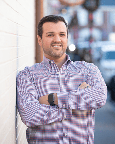 A man with short dark hair and a beard stands outdoors, smiling with arms crossed, leaning against a white brick wall. He wears a checkered button-down shirt and a smartwatch. The background shows a blurred street scene.