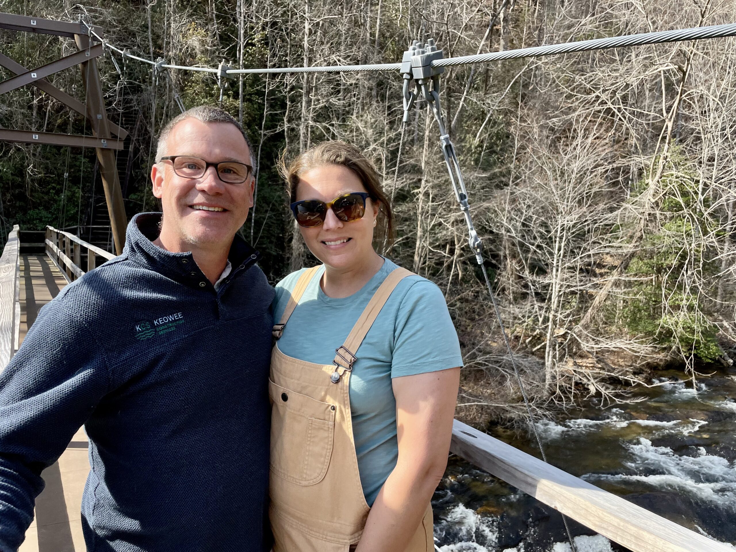 A man and woman smile while standing on a wooden bridge over a river, surrounded by trees with bare branches on a sunny day.