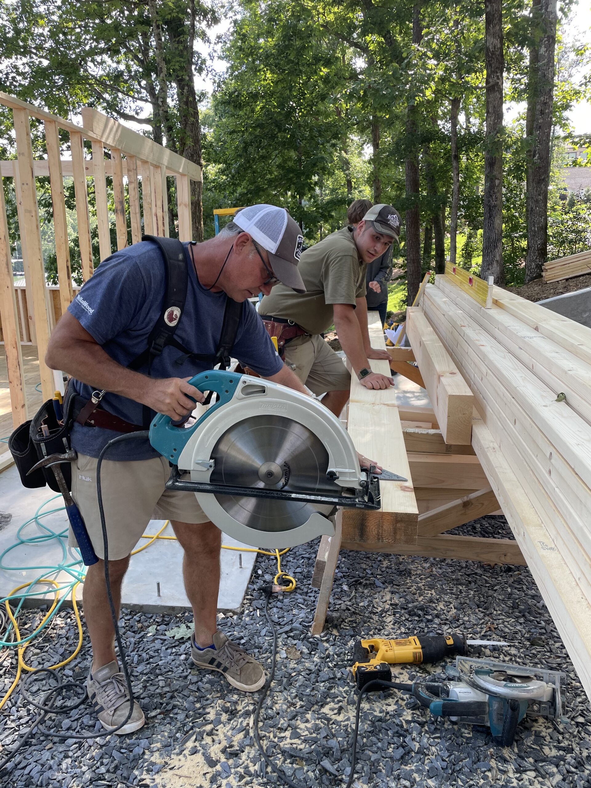 Two men work at a construction site outdoors. One is using a circular saw to cut wood, while the other measures lumber on a workbench. Framed walls and trees are visible in the background.
