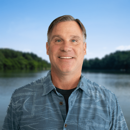 Middle-aged man with short brown hair wearing a blue patterned button-up shirt, smiling in front of a lake with trees and a clear blue sky in the background.