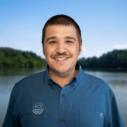 A smiling man with short brown hair and a mustache wears a blue button-down shirt with a "KCS" logo, standing in front of a scenic lake and trees under a clear sky.