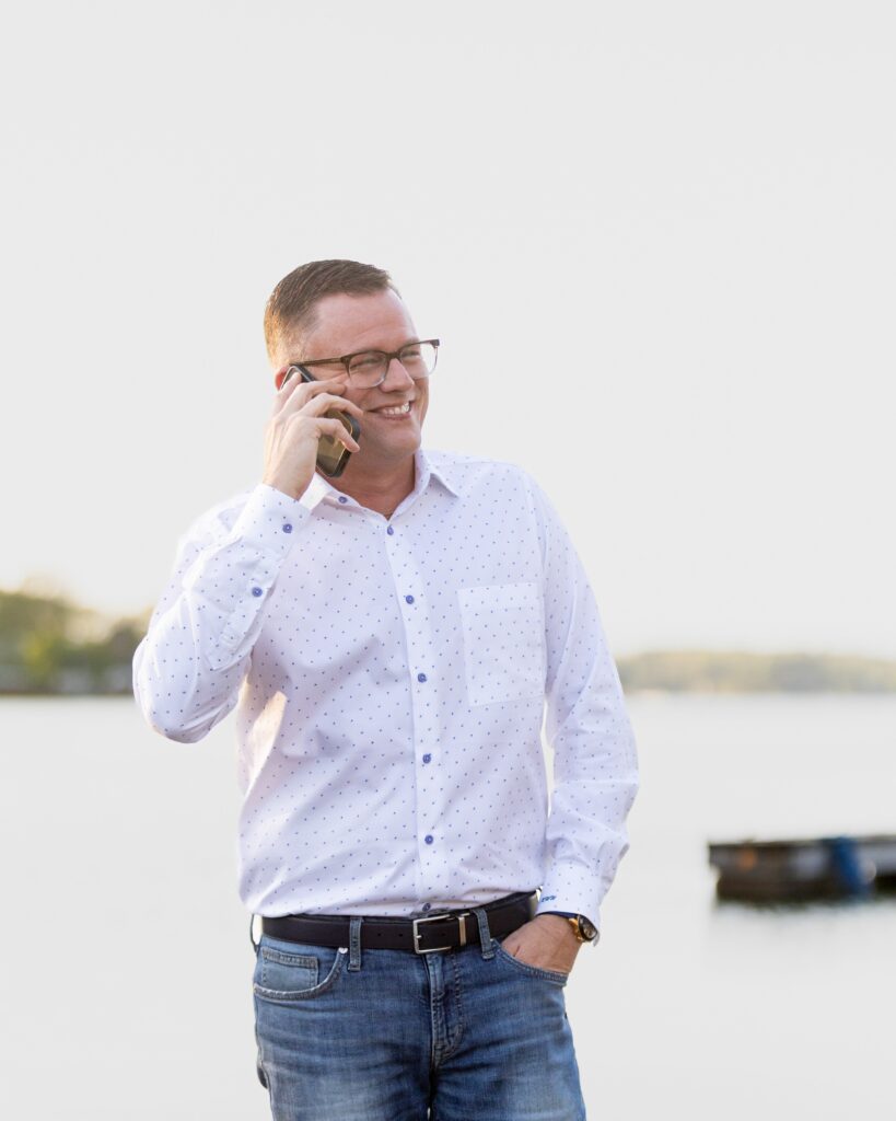 A man wearing glasses and a white button-down shirt smiles while talking on a cellphone, standing outdoors by a body of water with a blurred boat in the background.