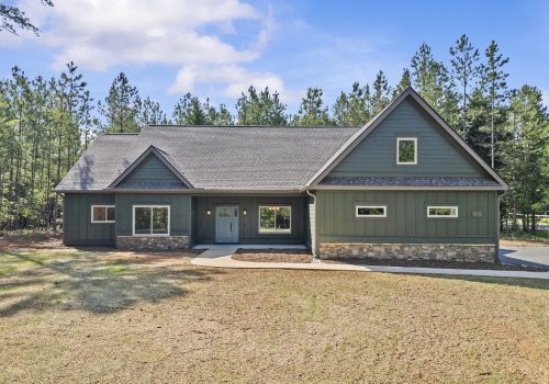 A modern green house with stone accents, gable roof, and a covered porch sits in front of a forest with tall pine trees under a blue sky with light clouds.