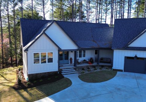 A modern white house with dark roofing, a covered front porch, two-car garage, and landscaped yard, surrounded by tall trees under a clear sky.