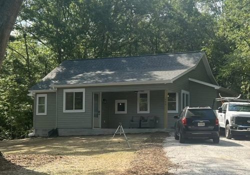 A small gray house with a covered front porch sits near trees. Two vehicles, an SUV and a truck, are parked on the driveway. The yard is patchy with grass and shaded by large trees.