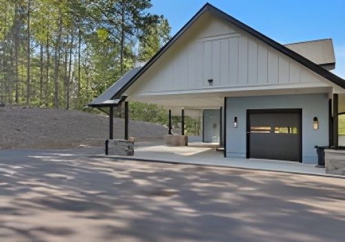 A modern, single-story building with a sloped roof and attached carport sits beside a paved driveway. Trees and greenery are visible in the background under a clear blue sky.