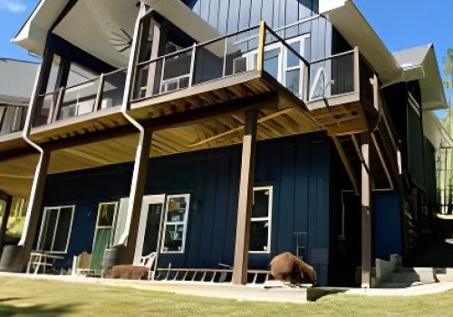 A modern two-story house with dark blue siding, large windows, a balcony with glass railing, and a covered patio area below, set on a neatly mowed lawn under a clear blue sky.