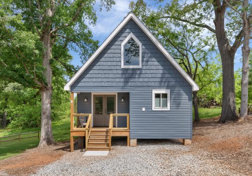 A small blue-gray house with white trim, a gabled roof, and a wooden front porch sits among tall green trees on a gravel driveway on a sunny day.