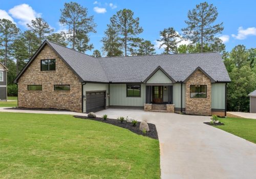 Modern single-story house with stone and green siding exterior, covered front porch, dark double garage, large driveway, and manicured lawn, surrounded by tall pine trees under a blue sky with clouds.