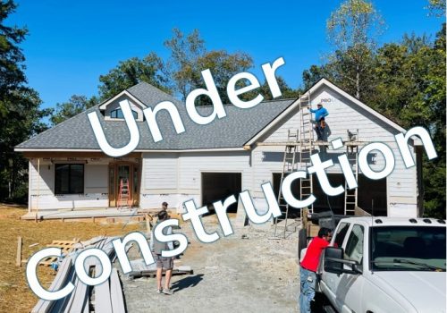 A house under construction with workers on scaffolding and near a white pickup truck, surrounded by trees. Large text across the image reads "Under Construction.