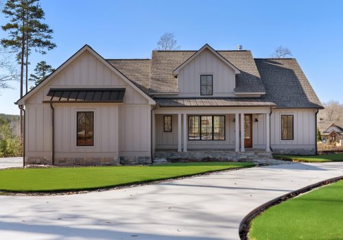 A modern, beige-colored house with a gabled roof, multiple large windows, and a wooden front door. The home features a well-maintained artificial lawn and a curved concrete driveway under a clear blue sky.