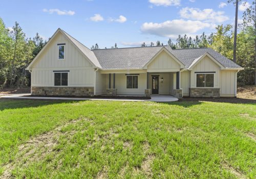 A modern single-story house with cream-colored siding, a gabled roof, stone accents along the base, large windows, and a spacious front yard with green grass, surrounded by tall trees under a blue sky with clouds.
